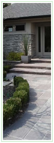 Curved stone walk with hedge leading to complete house exterior renovation in grey stone. Potscaping at front entrance.